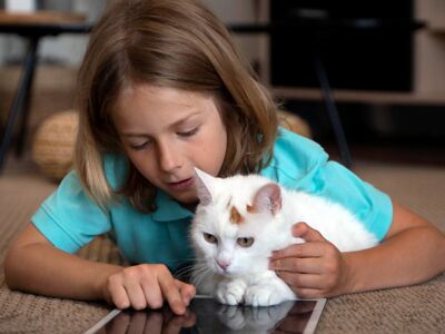 Ein Kind liegt mit einem Tablet auf dem Boden und hält eine weiße Katze im Arm.